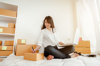 Smiling young woman with laptop writing on box while sitting in home office