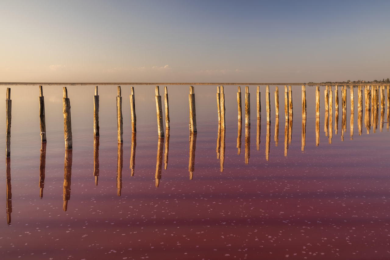 sky, horizon, reflection, water, nature, land, sea, tranquility, scenics - nature, morning, beauty in nature, no people, sunrise, wave, sunlight, dawn, beach, tranquil scene, horizon over water, wood, in a row, sand, clear sky, environment, outdoors, ocean, landscape, wooden post, idyllic, post, non-urban scene, shore, day