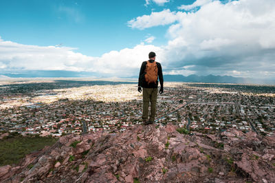 Rear view of male hiker standing on mountain with cityscape in background against cloudy sky