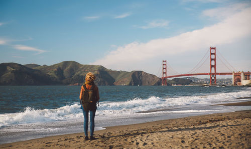 Rear view of woman standing on shore with golden gate bridge in background