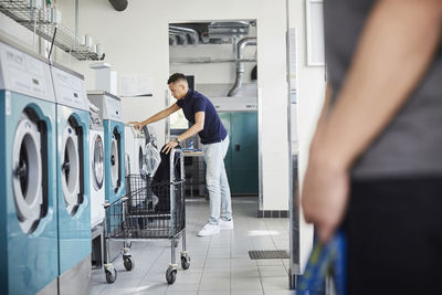 Full length of man using washing machine at laundromat with friend standing in foreground
