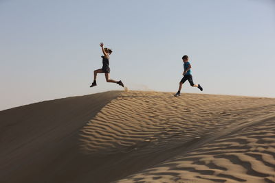 Siblings jumping at desert against clear sky