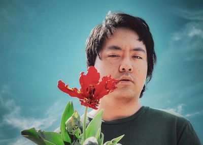 Low angle view of young asian man holding red tulip flowering plant against cloudy blue sky.