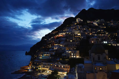 Illuminated buildings by sea against sky at dusk