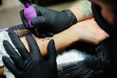 Unrecognizable concentrate bearded male master in black face mask and gloves creating tattoo on arm of crop faceless client in salon