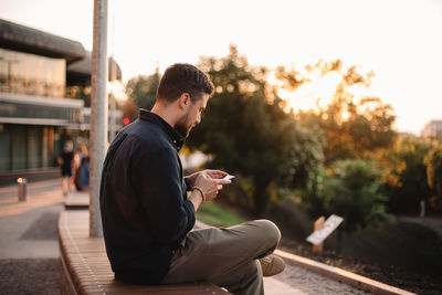 Happy man using smart phone sitting on bench at sunset