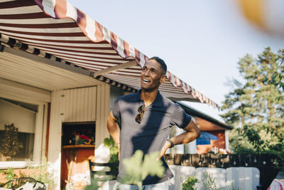 Happy young man looking away while standing with hands on hip at yard