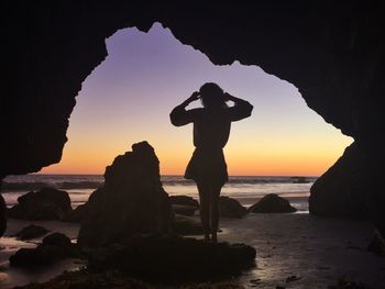 Rear view of silhouette woman standing at shore against sky during sunset