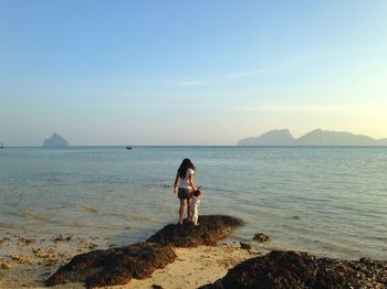 Full length of woman and baby standing on rock at beach against sky