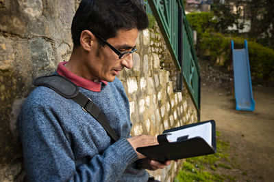 Man reading book while standing by stone wall