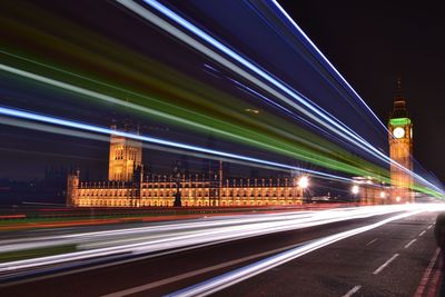 Light trails over street by big ben against sky at night
