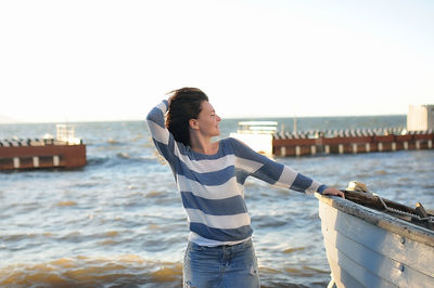 Nice girl curly hair in a striped sweater and a denim skirt with holes stands near an old gray boat