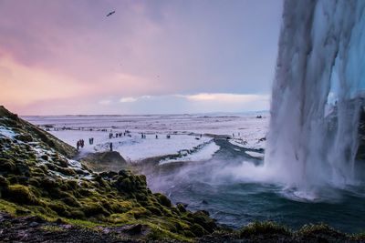 Scenic view of waterfall by snow covered landscape against cloudy sky during sunset