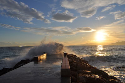 Waves splashing on pier against sky during sunset