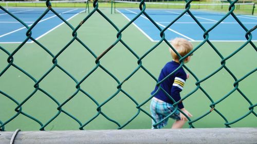 Rear view of boy running on playing field seen through chainlink fence
