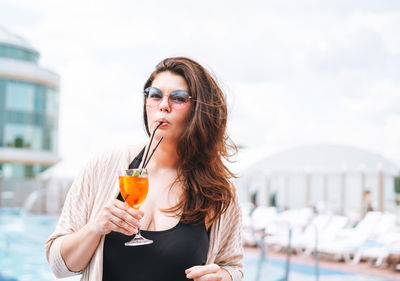 Stylish happy young woman in black swimsuit and sunglasses drinking cocktail near pool