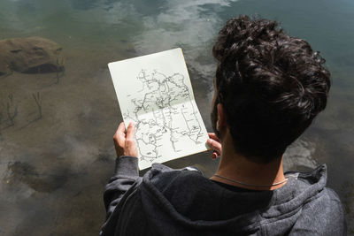 Back view of unrecognizable male explorer reading paper map and searching for route near pond in pyrenees mountain range