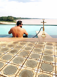 Rear view of shirtless man relaxing in swimming pool against sea