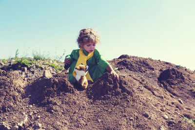 Boy sitting with stuffed toy on mud against sky