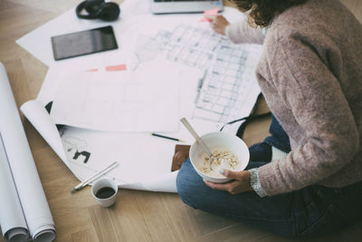 Woman working at home sitting on the floor looking at blueprint having breakfast
