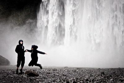 Young man holding hand of jumping girlfriend against waterfall