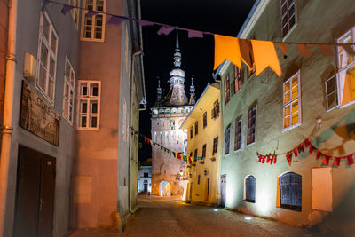 Narrow street amidst buildings in city at night. view of medieval clock tower in sighisoara, romania 