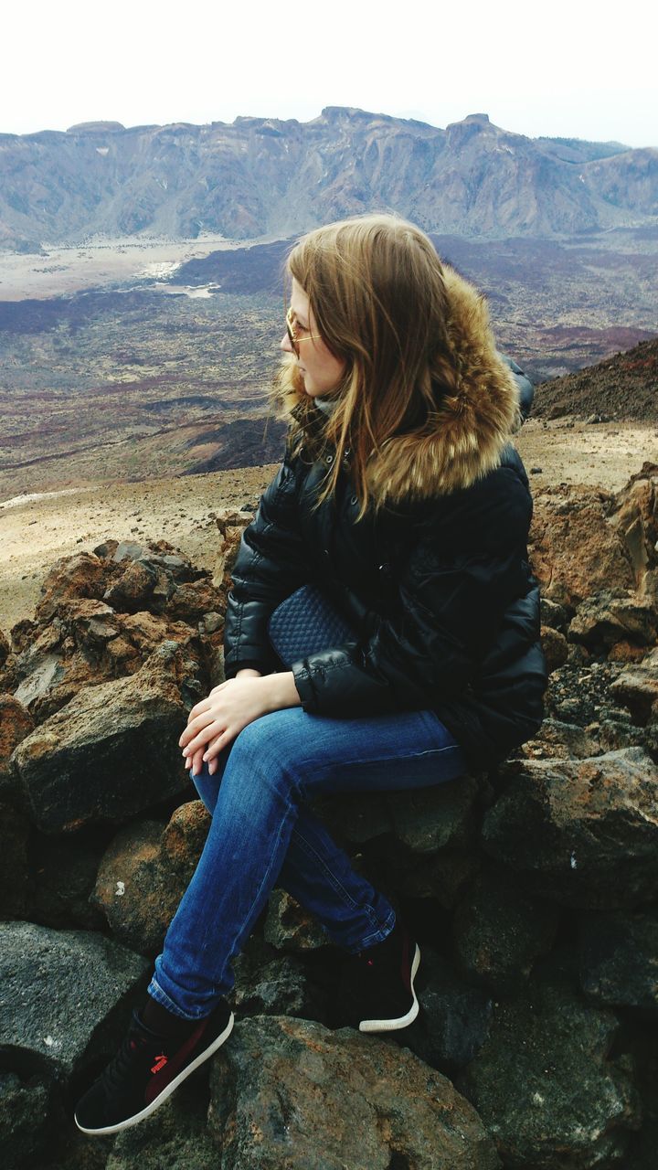 WOMAN SITTING ON ROCK AGAINST MOUNTAINS