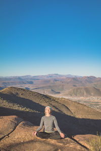 Woman practicing yoga on rock at mountain peak against blue sky during sunny day