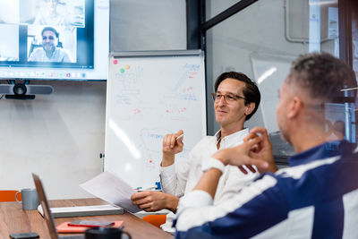 Coworkers sitting at table with gadgets and documents in modern workspace with white flip chat during online conference in office