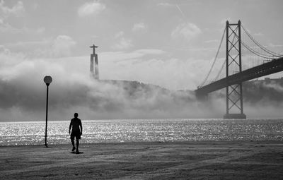 Silhouette man standing on street by tagus river and april 25th bridge against sky during foggy weather