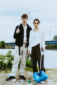 Full length portrait of young friends collecting plastic garbage near river against sky