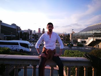 Portrait of young man sitting on cityscape against sky
