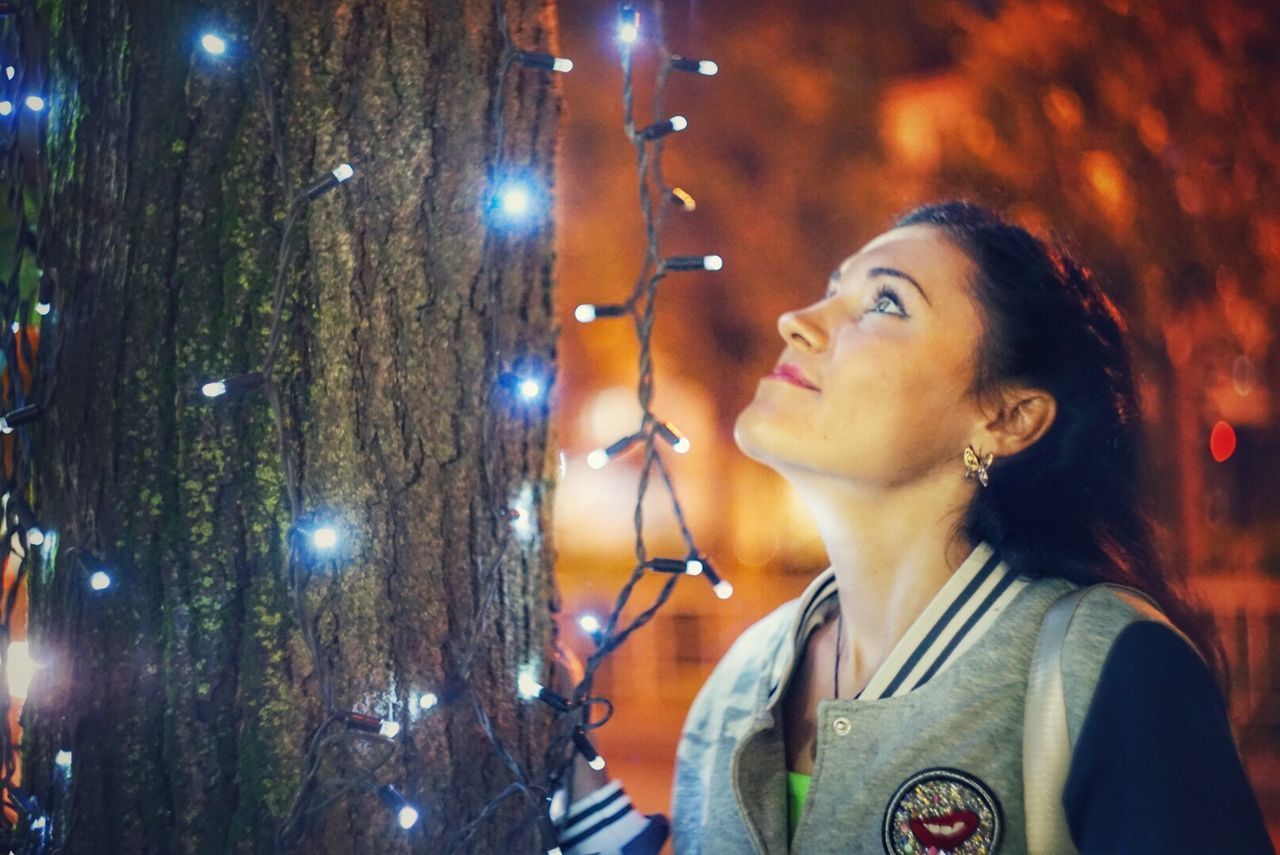 PORTRAIT OF YOUNG WOMAN LOOKING AT ILLUMINATED TREE TRUNK