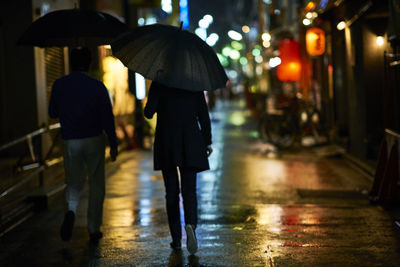 Rear view of people carrying umbrella while walking on wet street at night