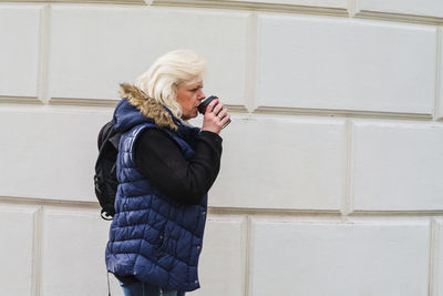 Side view of senior woman drinking coffee while standing against wall