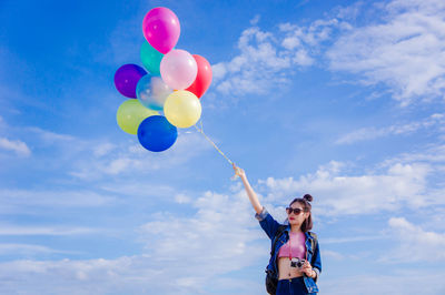 Young woman holding colorful balloons while standing against sky