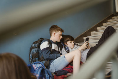 Male friends sharing smart phone while sitting on staircase of school building