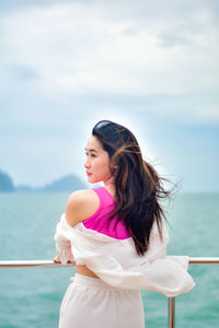 Side view of young woman with arms raised standing at beach against sky