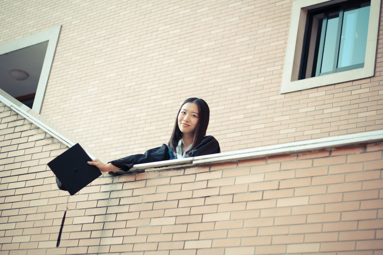 building exterior, architecture, lifestyles, built structure, young adult, casual clothing, leisure activity, person, wall - building feature, full length, front view, standing, young women, window, sitting, looking at camera, portrait