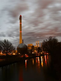 Illuminated factory by river against sky