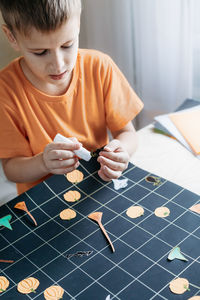 Cute child creating halloween-themed board game. boy gluing paper-cut game elements on playing field