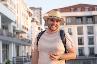 Handsome bearded smiling man,tourist, looking at camera,wearing sunhat, backpack, holding smartphone