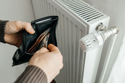 A man holds a wallet with banknotes of money inside next to a heating radiator. rising prices 