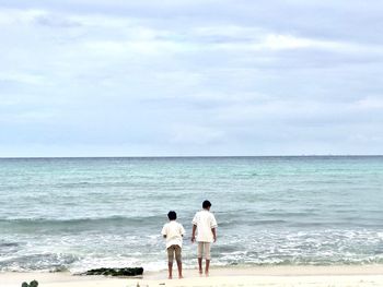 Rear view of male friends standing at beach against cloudy sky