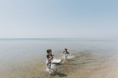 Brothers with surfboard running in sea against clear sky during sunny day