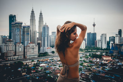 Rear view of woman with hands in hair standing by buildings against sky in city