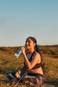 Smiling woman drinking water while sitting on grass against sky
