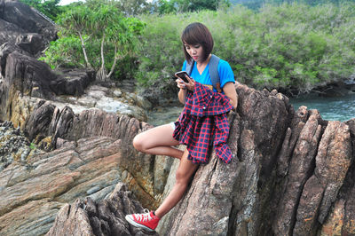 Young woman using mobile phone while leaning on rock formation against plants
