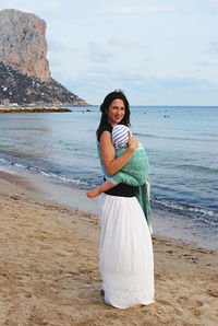 Young woman holding baby standing at beach against sky