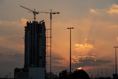 Low angle view of silhouette crane by building against sky during sunset